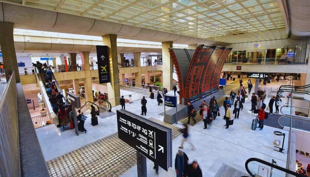 A bustling airport scene with passengers navigating the arrivals terminal at Paris Charles de Gaulle Airport. In the foreground, a group of travelers wheel their luggage through the expansive, well-lit space, their faces reflecting a mix of excitement and fatigue after a long flight. Rows of check-in counters and digital displays line the middle ground, providing information and directing the flow of traffic. In the background, floor-to-ceiling windows offer glimpses of the tarmac and aircraft, creating a sense of the larger transportation network. The image should convey the practical, efficient, and affordable options available for travelers to reach the city center from this major international hub. Saguão do Aeroporto Charles de Gaulle em Paris (CGD)