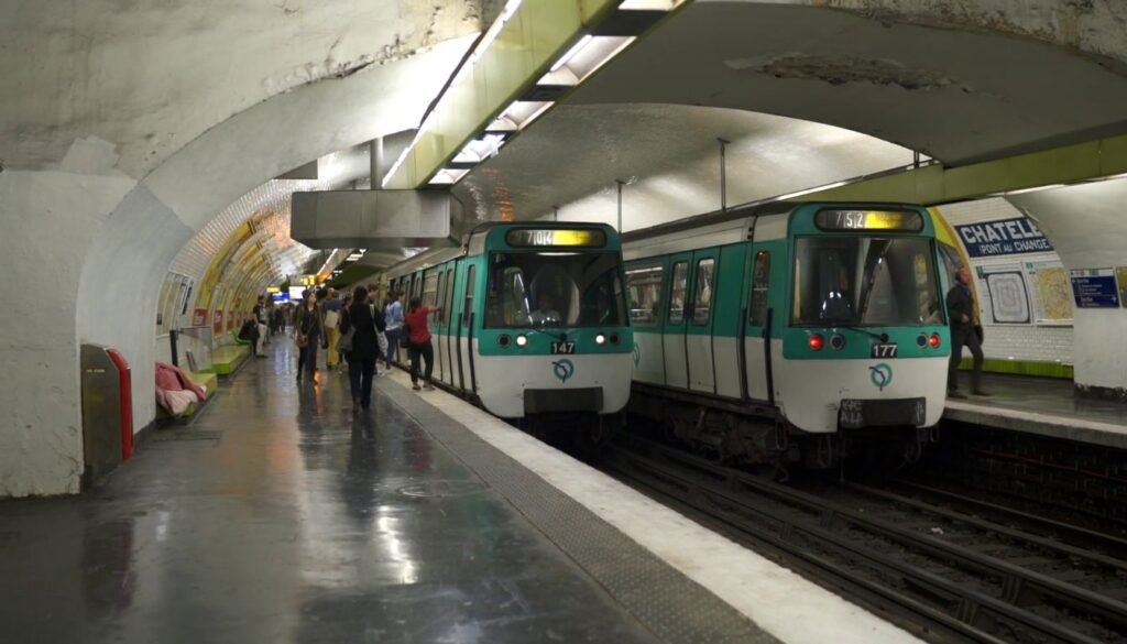 A bustling metro station in Paris, with a towering vaulted ceiling and gleaming marble floors. Commuters hurry along the platforms, their footsteps echoing through the cavernous space. Bright, natural light streams in through large windows, casting a warm glow over the scene. The station's art nouveau architectural details, such as ornate ironwork and intricate mosaics, lend an air of timeless elegance. In the background, a train slowly pulls into the station, its sleek, modern design a testament to the city's efficient public transportation network. The overall atmosphere conveys the vibrant, cosmopolitan nature of Paris, making this an ideal location for finding affordable yet well-situated hotels. Estação de Metrô em Paris.