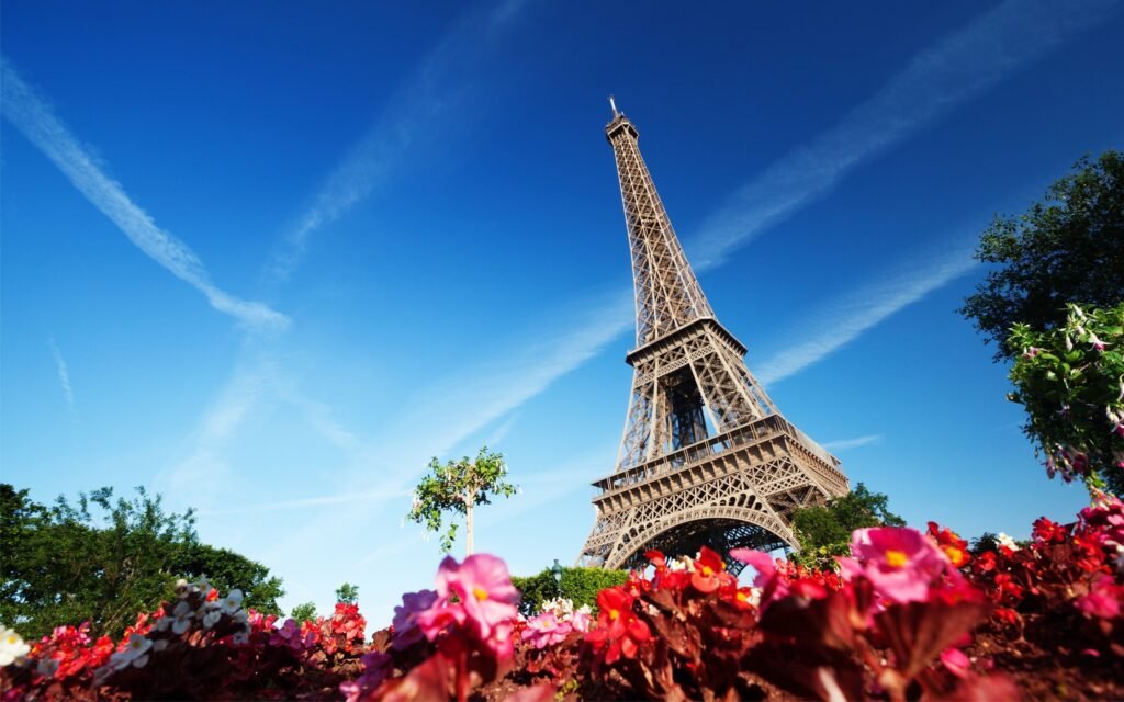 A vibrant springtime scene in Paris, France. In the foreground, a lush garden bursts with blooming flowers - rows of tulips, daffodils, and cherry blossoms swaying gently in a soft breeze. The middle ground reveals a picturesque cobblestone street, flanked by charming Parisian townhouses and quaint cafés with wrought-iron tables and bistro chairs. In the background, the iconic Eiffel Tower stands tall, bathed in warm, golden-hour light. Pedestrians stroll leisurely, enjoying the mild weather and lack of crowds. The overall atmosphere is one of tranquility, romance, and the joyful energy of springtime in the city of light. Primavera em Paris. Flores em torno da Torre Eiffel