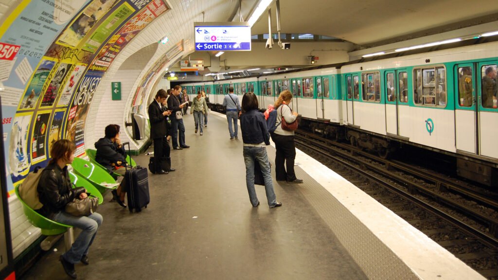 A bustling Parisian metro station, with sleek and modern trains gliding in and out of the platform. Bright overhead lighting illuminates the scene, casting warm reflections on the polished tile floors. Passengers hurry along the walkways, their faces a blend of purpose and anticipation. In the background, ornate architectural details adorn the vaulted ceilings, hinting at the station's historic roots. The atmosphere is one of efficiency and urban energy, perfectly capturing the essence of Paris's renowned public transportation system. O Metrô de Paris é uma ótima forma de economizar Tempo e Dinheiro.