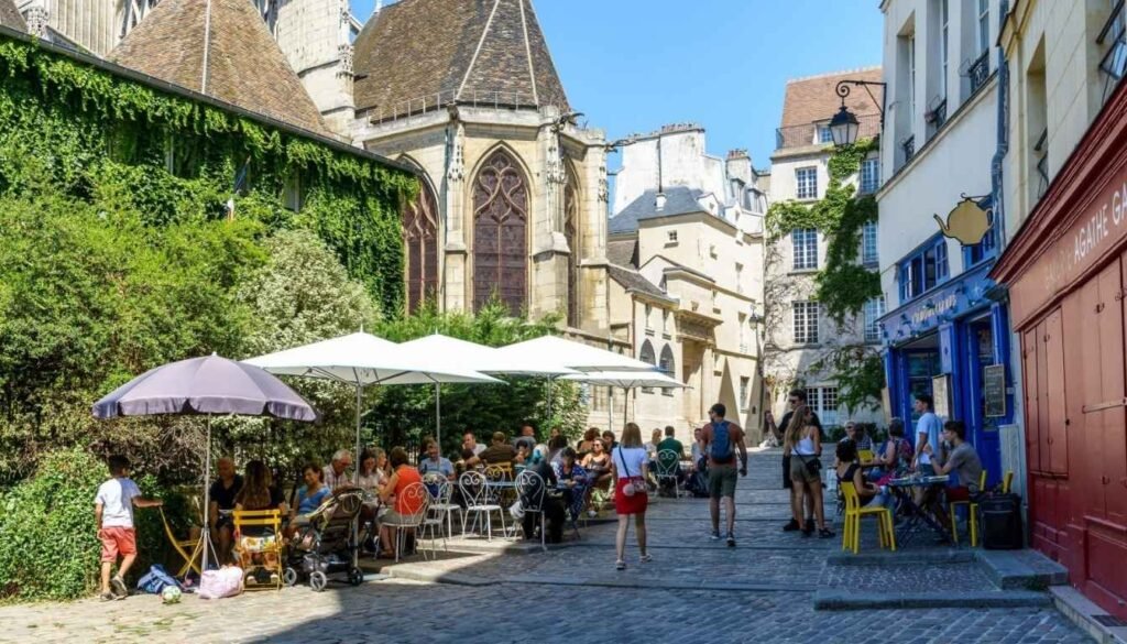 A bustling Parisian street scene at golden hour, with rows of charming townhouses lining the sidewalks. In the foreground, people stroll leisurely, café tables spill onto the pavement, and the iconic Métro station entrance is visible. The middle ground features a tree-lined boulevard, with the distant silhouette of the Eiffel Tower peeking through. The warm, soft lighting casts a romantic glow, evoking the essence of Parisian hospitality and the perfect setting to explore the city's accommodations. Cena no bairro Le Marais