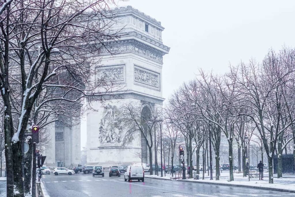 A serene winter scene in Paris, France. A snow-dusted cityscape with iconic landmarks like the Eiffel Tower and Notre-Dame Cathedral visible in the distance, their silhouettes emerging through a soft, hazy mist. In the foreground, a quaint cobblestone street lined with charming Parisian cafes and boutiques, their warm yellow lights casting a cozy glow onto the tranquil, empty street. Bare tree branches sway gently in the crisp air, and a light snowfall adds to the peaceful, romantic atmosphere. The scene is captured through a medium-wide angle lens, using cool, muted tones to evoke the charm and economy of the Parisian winter. Paris no Inverno