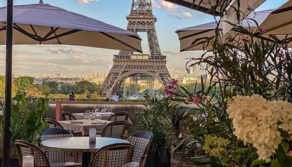 A charming Parisian street in the off-season, bathed in soft, golden light. Cobblestone paths wind through a picturesque neighborhood, lined with quaint cafes, boulangeries, and small boutiques. Locals stroll leisurely, sipping cafe au lait and admiring the elegant 19th-century architecture. In the distance, the iconic silhouette of the Eiffel Tower rises, its wrought-iron lattice framed by a clear, hazy sky. The scene exudes a sense of tranquility and joie de vivre, inviting the viewer to slow down and savor the understated beauty of Paris in the low season. Aproveite os restaurantes menos lotados em Paris durante a baixa temporada