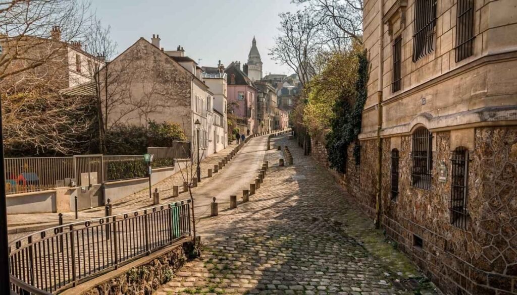 A picturesque Parisian street scene in the early morning light. The historic architecture of the city's iconic buildings stand tall and proud, their ornate facades casting long shadows across the empty cobblestone roads. A few lone pedestrians stroll leisurely, taking in the tranquil ambiance devoid of the usual bustling crowds. Warm, muted tones create a sense of timeless elegance, while a shallow depth of field focuses the viewer's attention on the serene, people-free foreground. The overall mood is one of peaceful solitude, inviting the viewer to imagine exploring Paris during its most pleasant, off-peak season. Rua em Montmartre