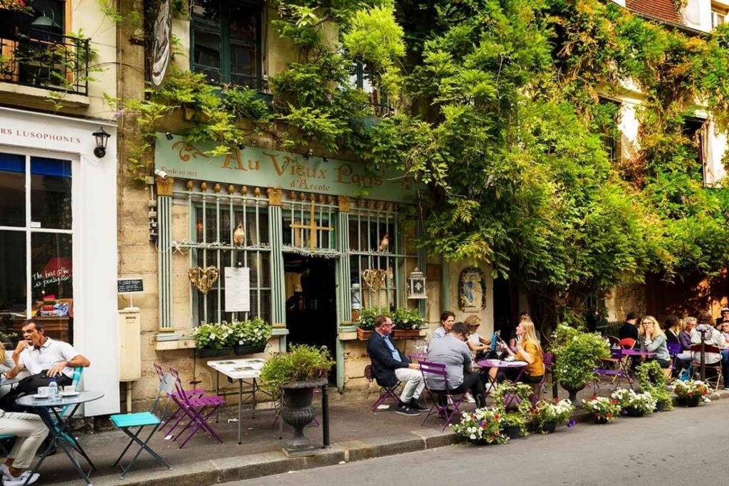 A bustling Parisian sidewalk cafe, sunlight spilling across the intricate wrought-iron tables and chairs. In the foreground, patrons savor delicate crepes and sip rich espresso, engaged in lively conversation. The middle ground features a charming storefront, its window displays tempting an array of artisanal breads and pastries. The background frames the iconic Eiffel Tower, its silhouette hazy in the distance, while the sky overhead is tinged with a soft, golden glow. The scene evokes the quintessential Parisian gastronomic experience - a harmonious blend of flavors, culture, and timeless ambiance. Um Bistrô em Paris