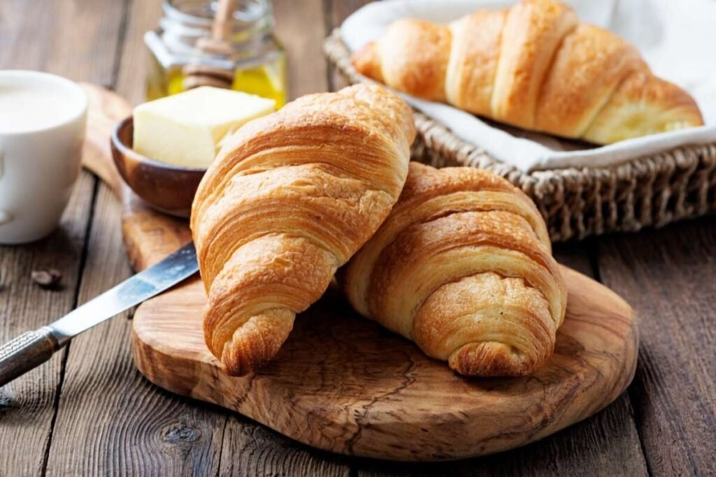 A freshly baked, golden-brown croissant sits on a delicate porcelain plate, casting a warm, inviting glow under the soft, diffused lighting of a Parisian café. The flaky, buttery pastry is perfectly shaped, with distinct layers that beckon to be torn apart and savored. The croissant is accompanied by a small dish of rich, creamy butter and a steaming cup of fragrant, aromatic coffee, creating a quintessential Parisian breakfast scene. The overall composition evokes the charm and elegance of the Parisian culinary experience, reflecting the section title "Minhas comidas típica de paris favoritas". Croissat e Manteiga, tipicamente parisienses