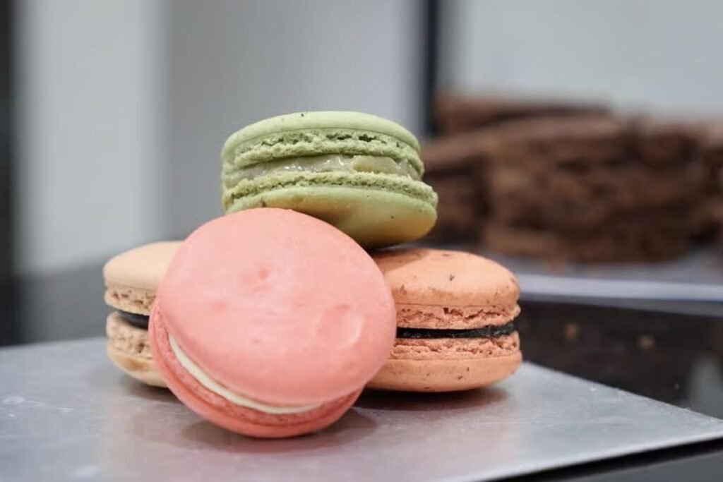 A delightful arrangement of delicate French macarons, meticulously crafted with vibrant pastel hues and elegant shapes. The macarons are beautifully displayed on a rustic wooden table, bathed in soft, natural lighting that casts gentle shadows and highlights their intricate textures. In the background, a blurred Parisian cityscape serves as a subtle nod to the Parisian origin of this beloved confection. The overall scene exudes a sense of whimsical sophistication, perfectly capturing the essence of the "Doces e sobremesas que viraram lembrança de viagem" section of the article. Macarons, doces típicos da Culinária francesa