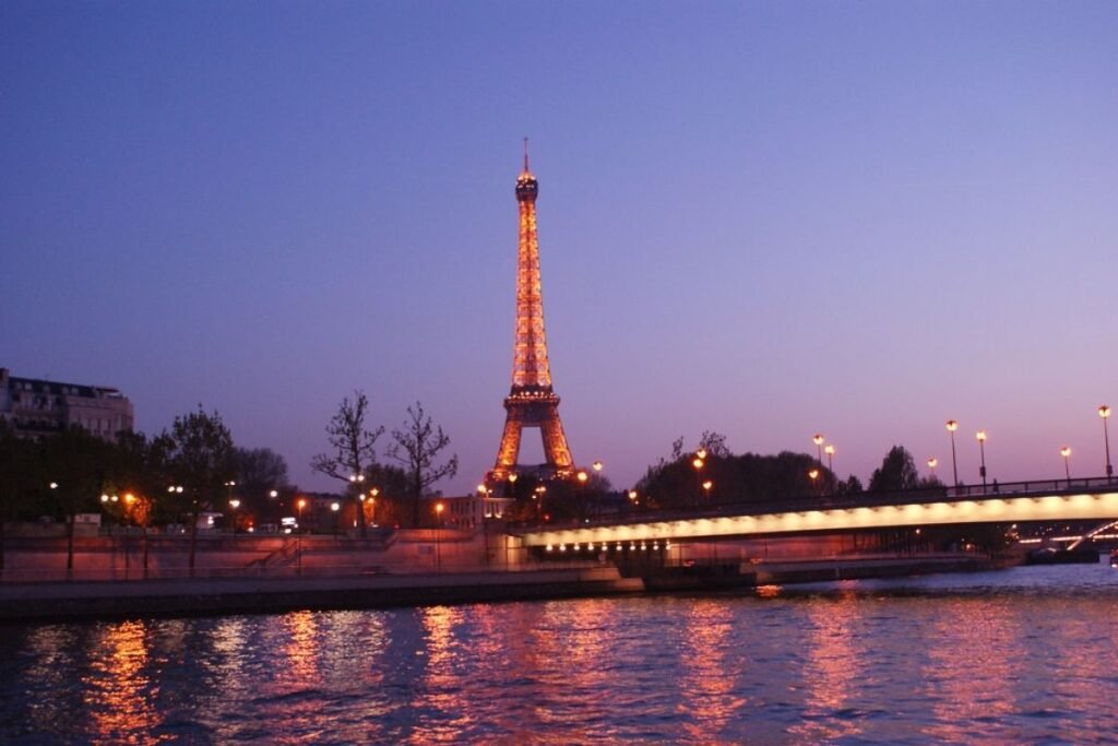 A romantic Parisian cityscape with the iconic Eiffel Tower as the central focus. Warm, golden hour lighting illuminates the city's grand architecture and cobblestone streets, creating a dreamlike atmosphere. In the foreground, a picturesque Parisian café with wrought-iron chairs and a striped awning sets the scene. In the middle ground, pedestrians stroll along the Seine River, taking in the stunning views. The background is filled with the charming rooftops, spires, and domes that make Paris the "City of Light." The overall composition conveys the timeless elegance and allure of this world-renowned destination. A Torre Eiffel iluminada ao entardecer e as luzes refletidas no Rio Sena